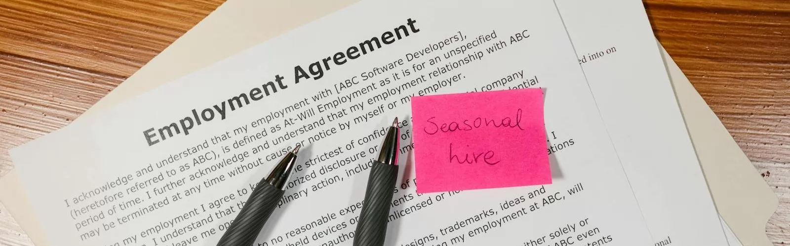 Top view of employment agreement contract with pens and seasonal hire note on wooden desk.